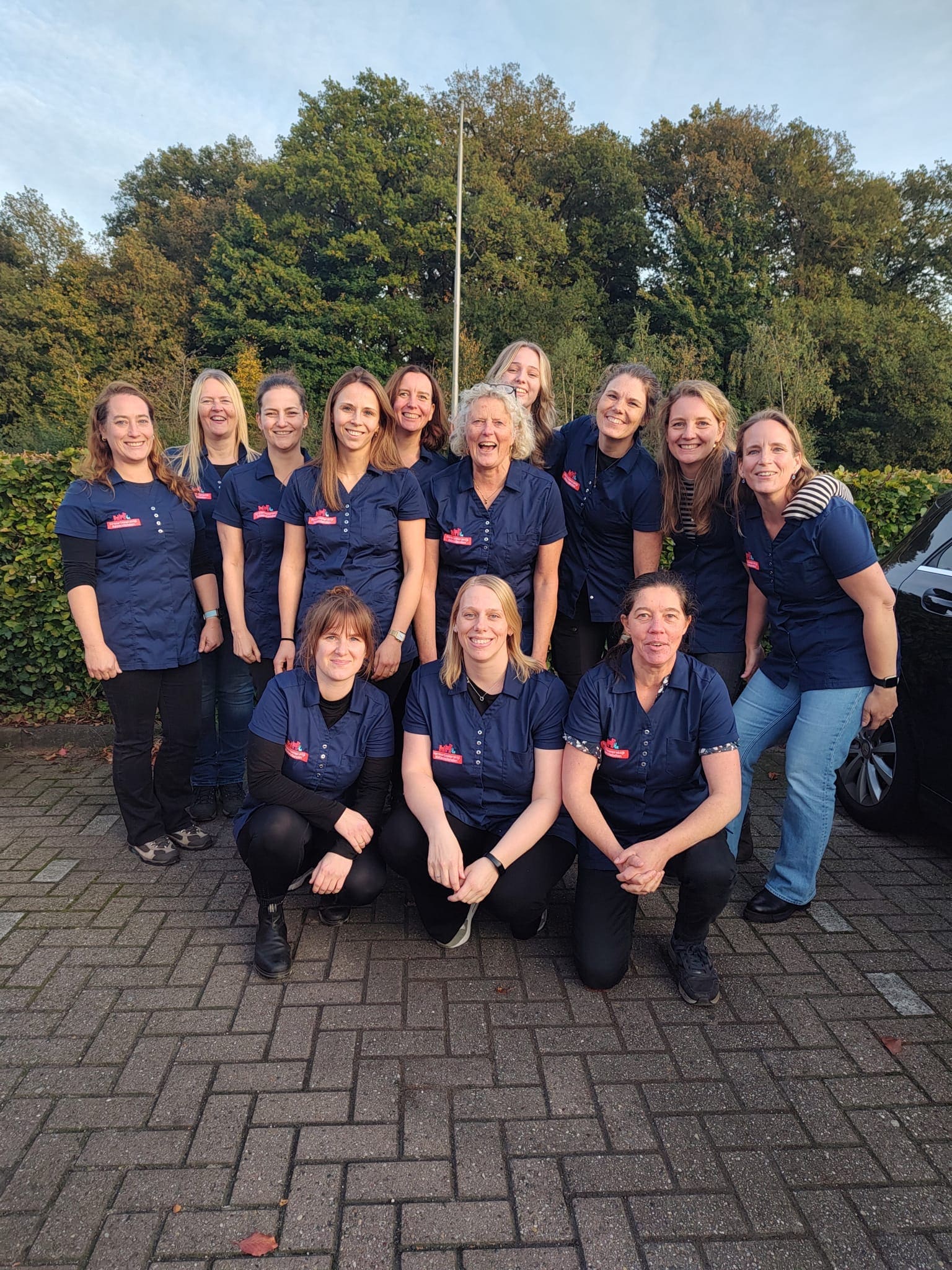 Het team van Dierenartspraktijk Keizerslanden in donkerblauwe praktijkshirts, samen lachend op een terrein voor hoge bomen